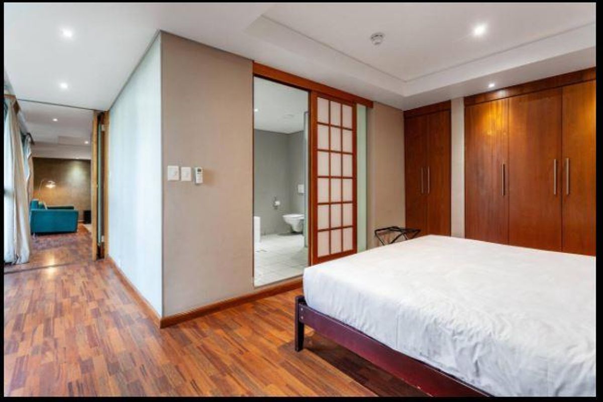 Spacious bedroom featuring warm-toned laminate flooring, extensive built-in wooden cabinetry, recessed lighting, and a sliding door leading to an en-suite bathroom.