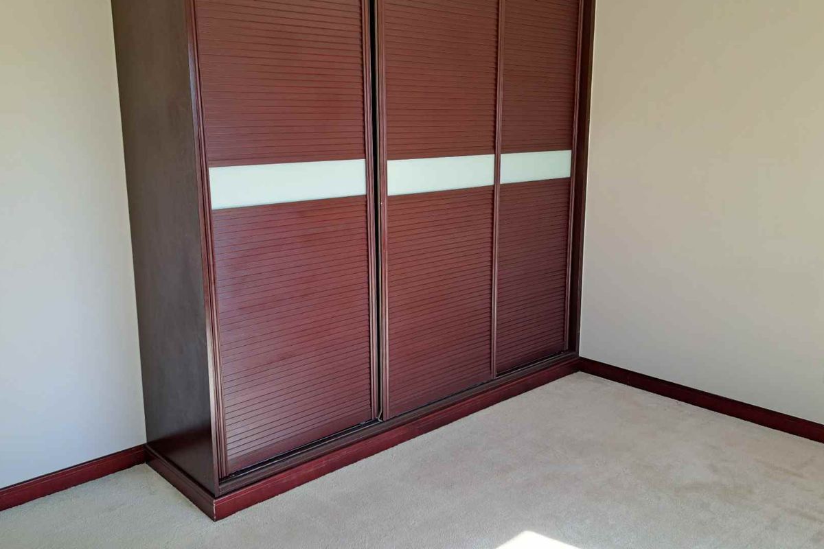 A bedroom featuring a large, built-in dark wood wardrobe with sliding doors and integrated frosted glass panels, set against neutral walls and carpeted flooring.