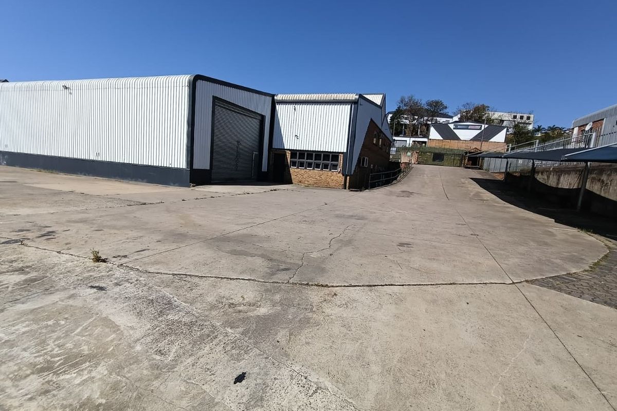 Industrial property exterior featuring a large concrete yard, corrugated metal warehouse structure, roller shutter door, and covered parking area.