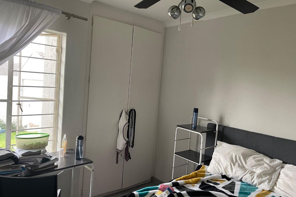 A bedroom featuring built-in cupboards, a ceiling fan, and a window with security bars, providing a functional living space.