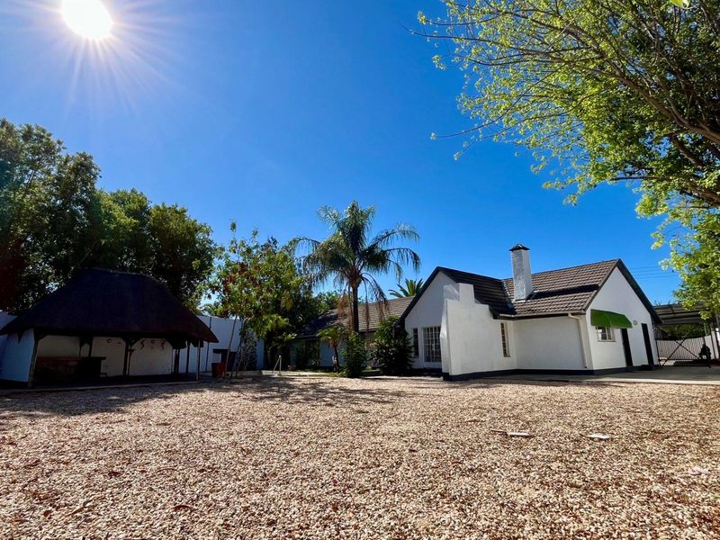 Spacious rear exterior area featuring a gravel yard, a traditional thatched lapa, and a well-maintained white building with a tiled roof.