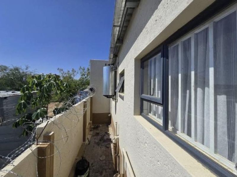 A narrow exterior side passage featuring a solar geyser, paved flooring, and security boundary wall with razor wire.