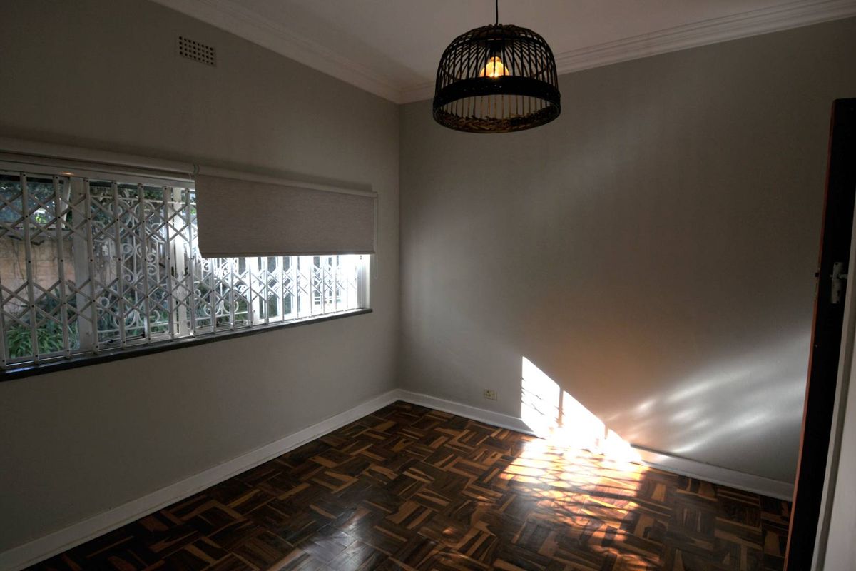 A bright, empty bedroom featuring polished parquet flooring, neutral wall tones, and a secure window with fitted burglar bars.