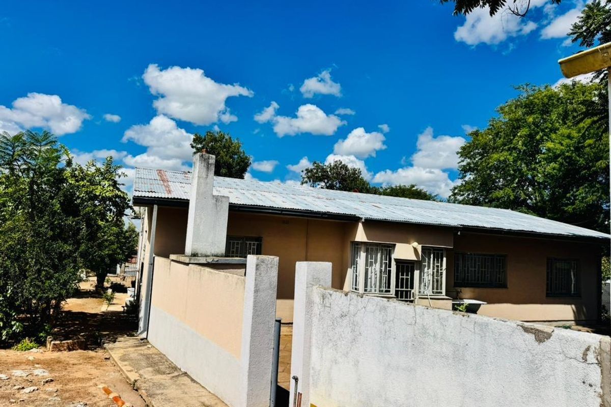 Exterior view of a single-story house featuring a corrugated metal roof, masonry boundary walls, and security bars on windows.