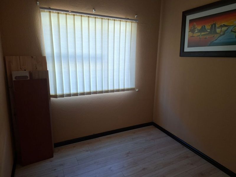 A simple, empty bedroom featuring light-toned laminate flooring, dark skirting boards, and vertical window blinds.