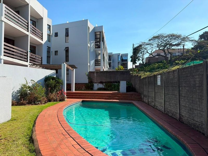 Communal swimming pool area featuring brick paving, a pergola, and surrounding apartment building balconies.