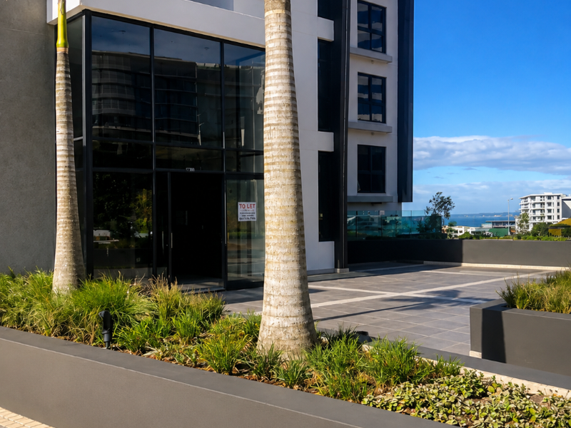 Modern commercial building entrance featuring a glass facade, paved terrace area, and landscaped raised planters.