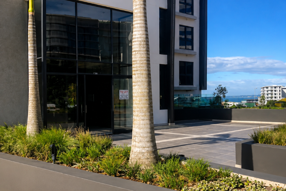 Modern commercial building entrance featuring a glass facade, paved terrace area, and landscaped raised planters.
