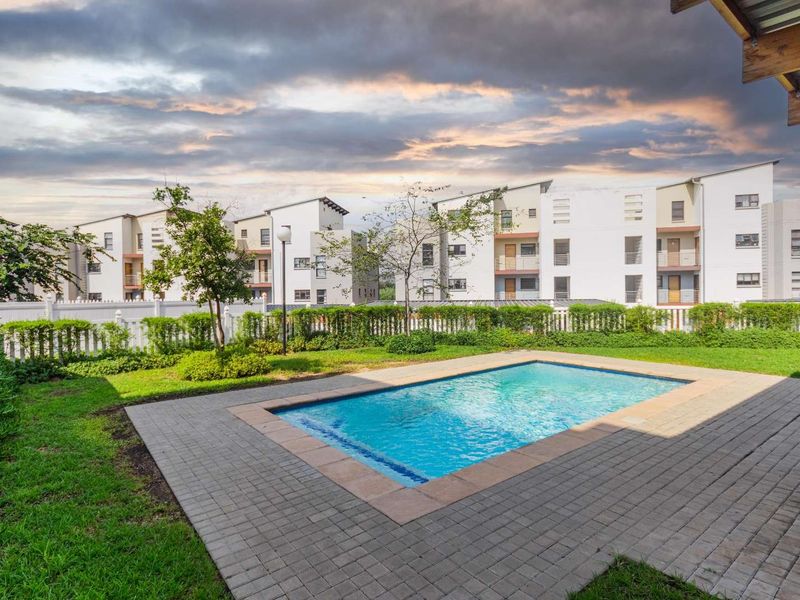A well-maintained communal swimming pool area featuring a paved deck, landscaped garden, and white picket fencing within an apartment complex.