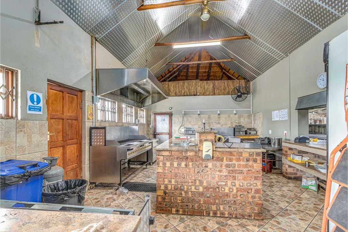 Commercial kitchen featuring a central brick island, stainless steel extractor hood, tiled splashbacks, and high vaulted ceiling with exposed metal roofing.