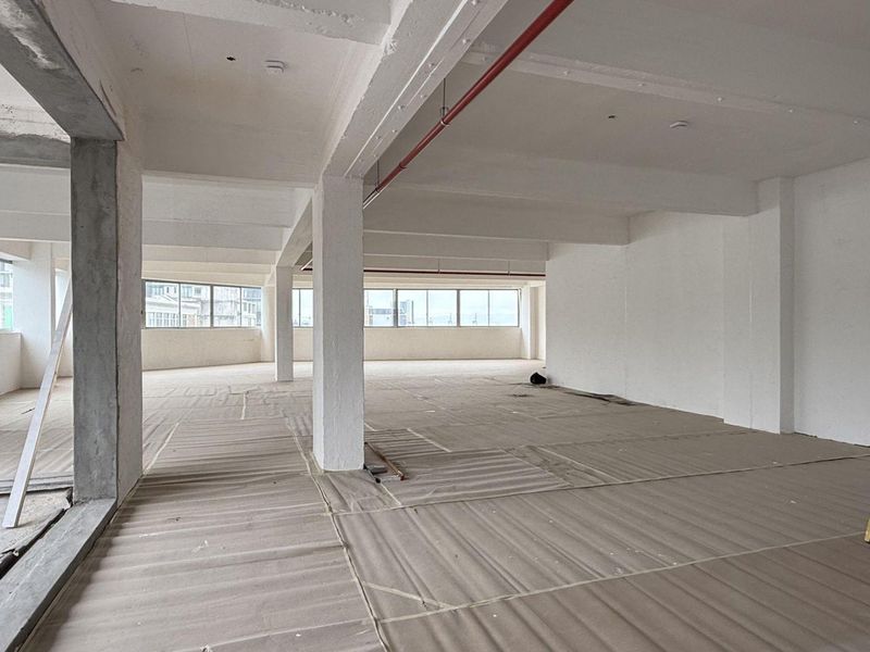 An expansive, open-plan office space currently under renovation, featuring exposed structural columns, high ceilings with fire suppression systems, and large windows providing natural light.