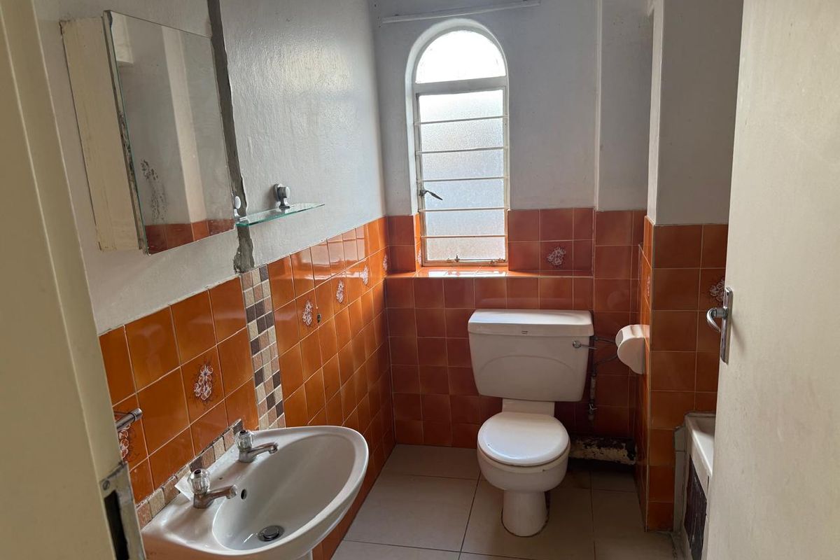 A dated bathroom featuring orange ceramic wall tiles, a pedestal basin, and a toilet, with an arched window providing natural light.