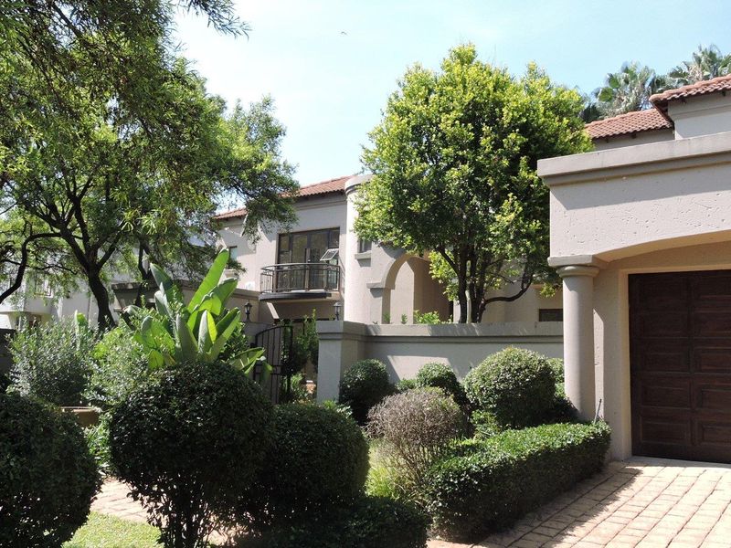Front elevation of a residential property featuring a paved driveway, garage, and established garden with mature trees and shrubs.