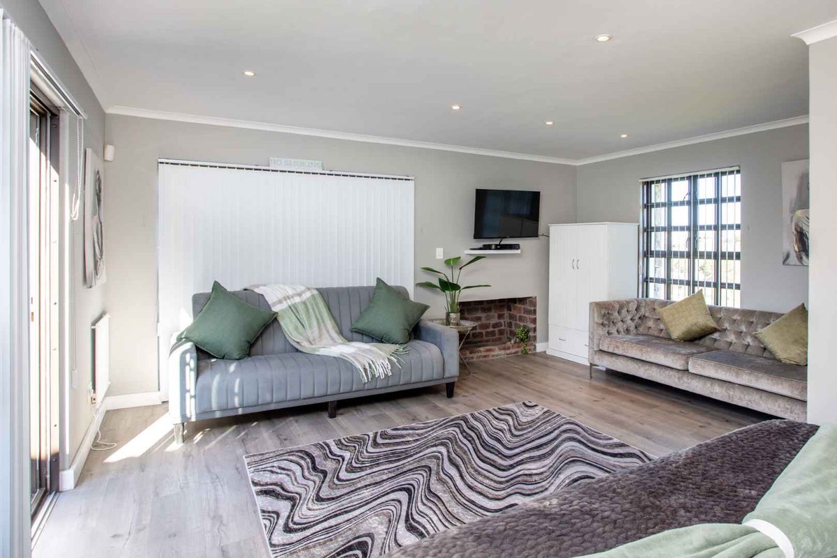 A spacious living room featuring light wood-look flooring, recessed downlighting, and a brick-accented fireplace area with large windows providing natural light.