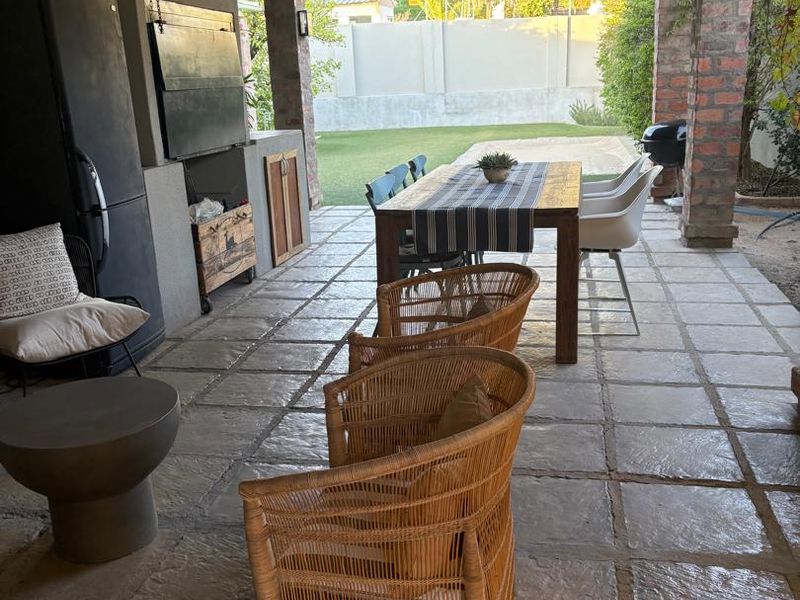 A spacious covered patio featuring stone tile flooring, exposed brick pillars, and a built-in braai area, overlooking the rear garden.