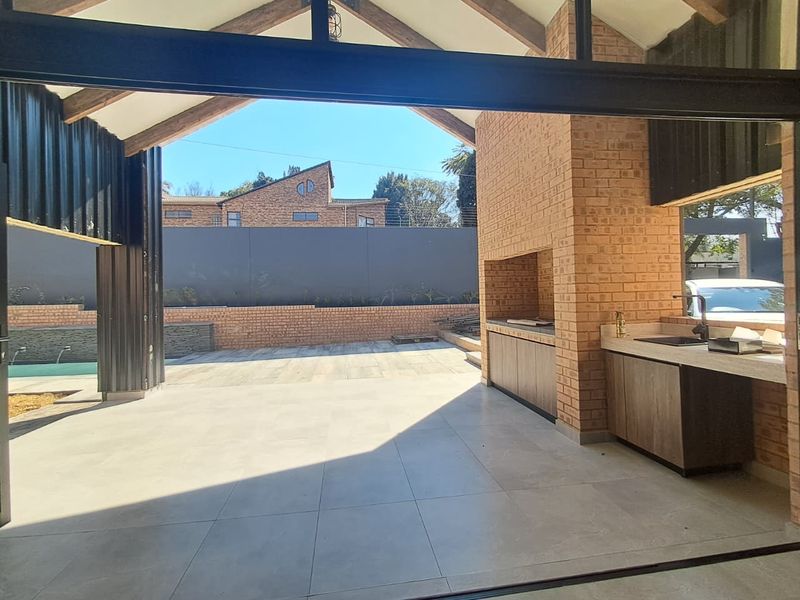 Spacious covered patio featuring exposed timber trusses, a built-in braai, and a functional prep area with cabinetry and a sink, overlooking the garden.