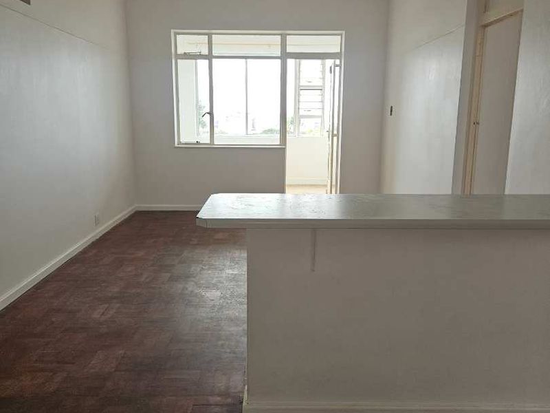 An empty open-plan living area featuring original parquet flooring, a breakfast bar, and natural light from a large window leading to a balcony.