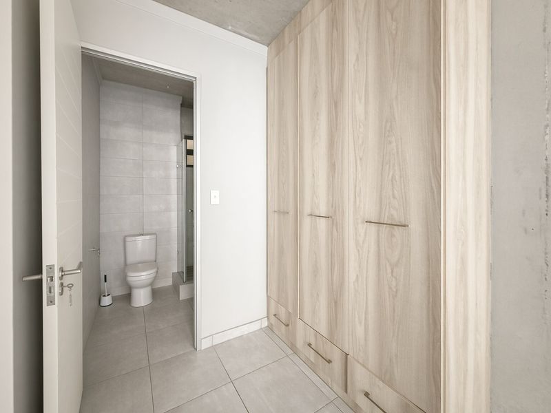 Modern bedroom featuring built-in light wood cupboards, tiled flooring, an exposed concrete ceiling, and an adjoining en-suite bathroom.