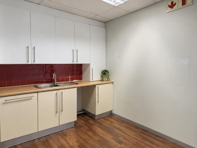 A clean, functional office kitchenette featuring white cabinetry, a wood-look laminate countertop, a stainless steel sink, and a red tiled backsplash.