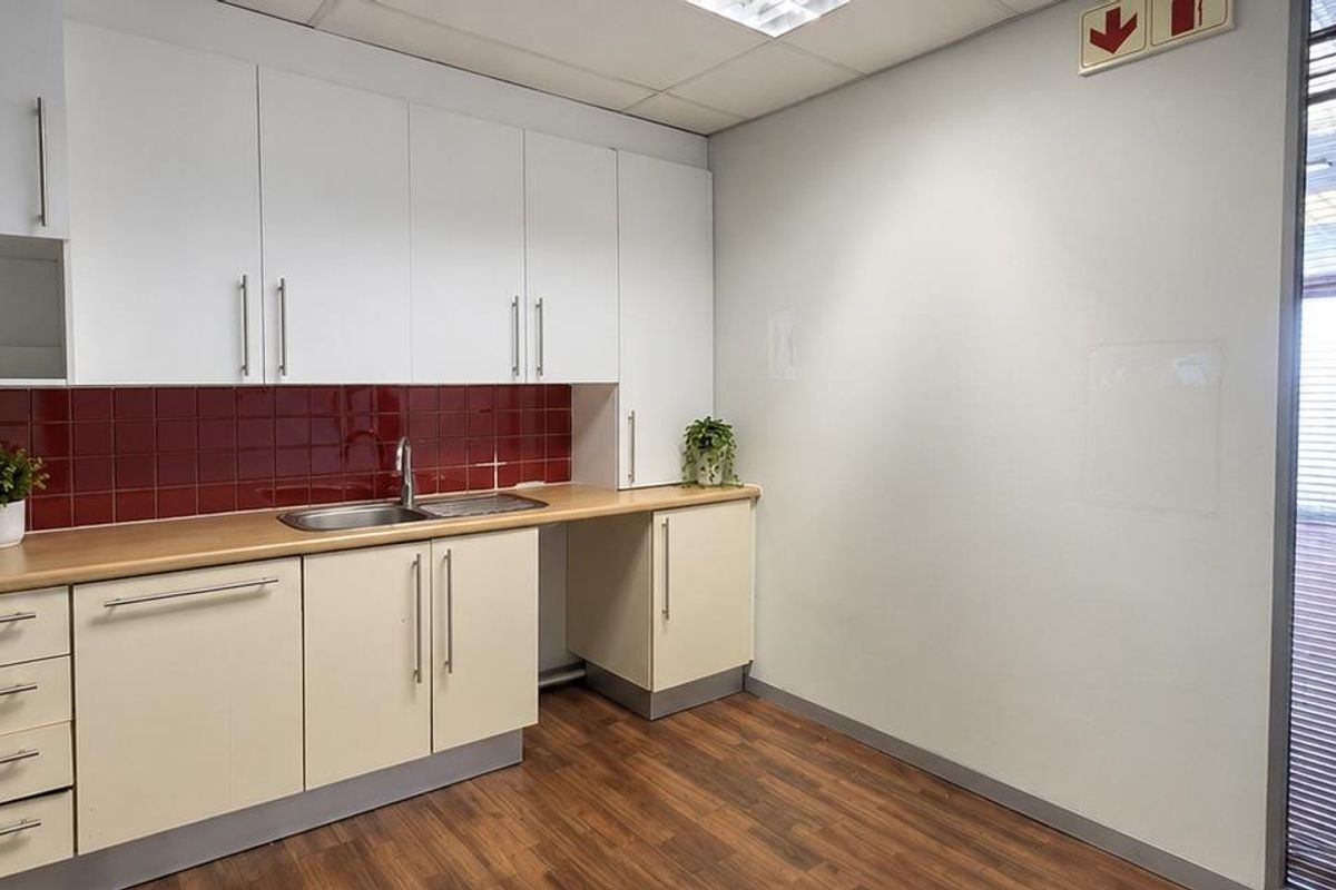 A clean, functional office kitchenette featuring white cabinetry, a wood-look laminate countertop, a stainless steel sink, and a red tiled backsplash.