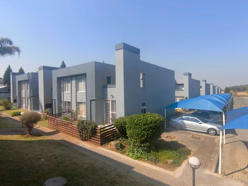 Exterior view of a modern townhouse complex featuring paved driveways, covered carport parking with shade sails, and well-maintained landscaped garden areas.