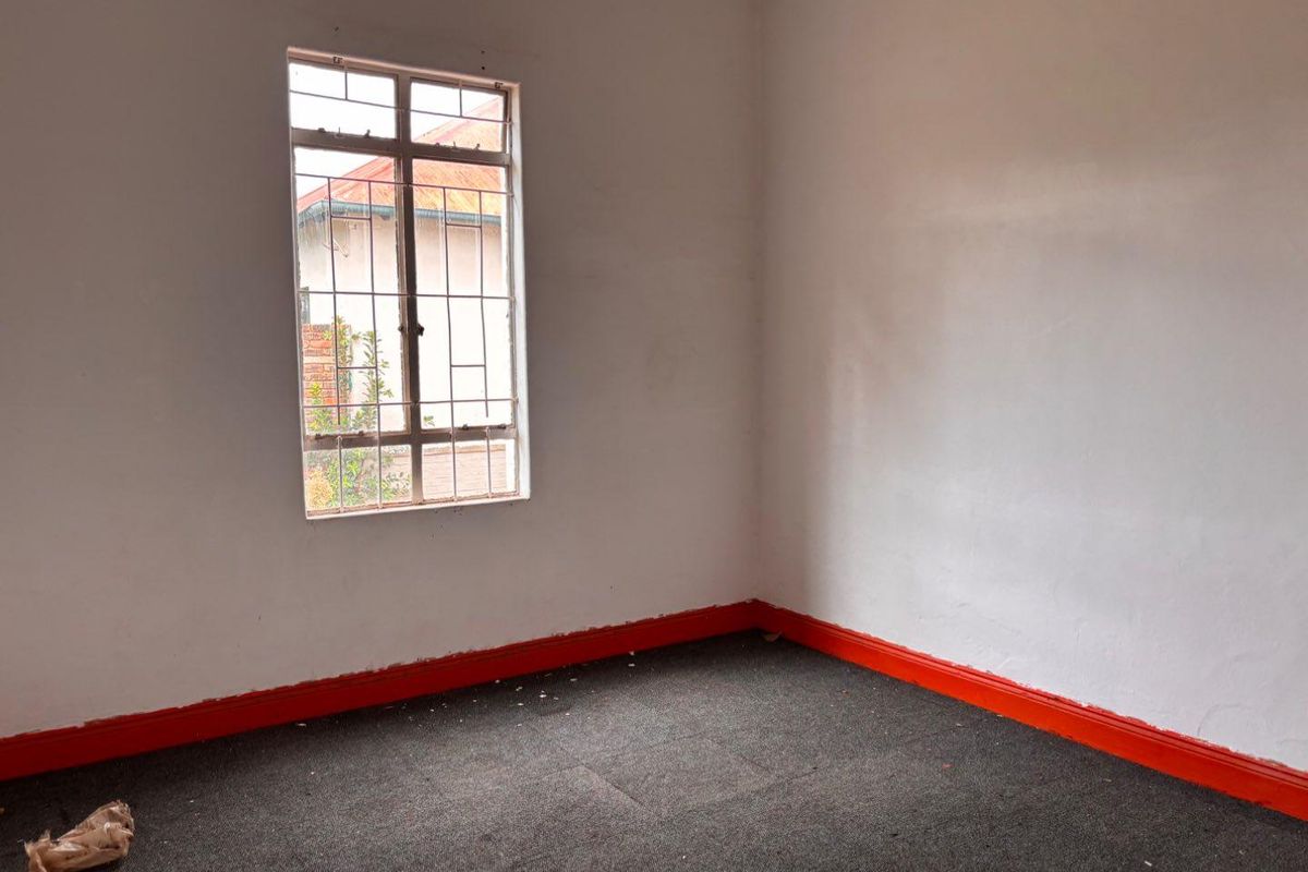 A vacant bedroom featuring grey carpeting, bright red skirting boards, and a window with integrated security bars.