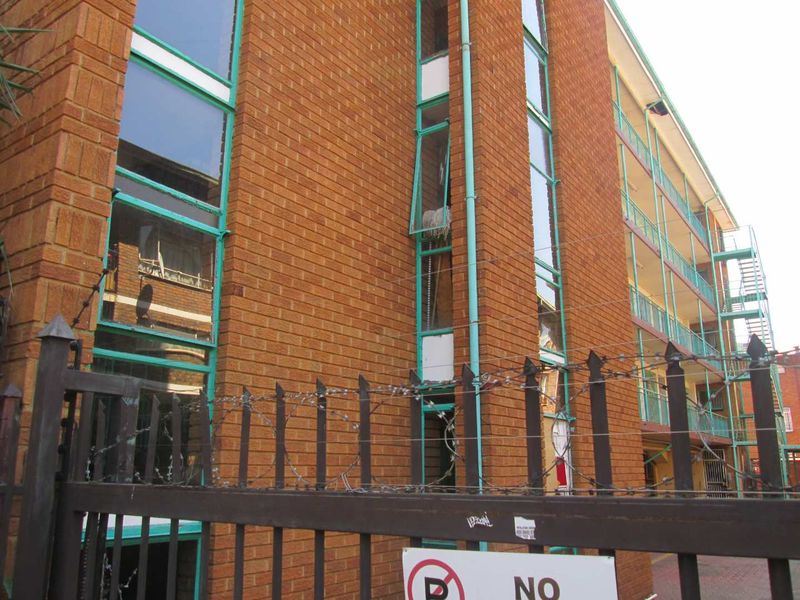 Exterior view of a multi-story apartment building featuring brick facade, metal window frames, and perimeter security fencing with barbed wire.