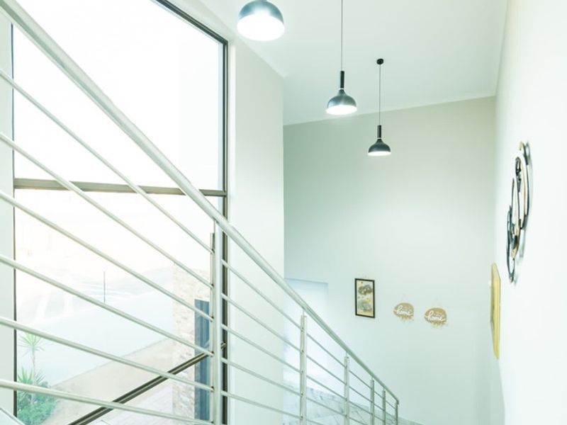 A bright and modern staircase area featuring tiled flooring, a sleek stainless steel balustrade, and contemporary pendant lighting with a large window.