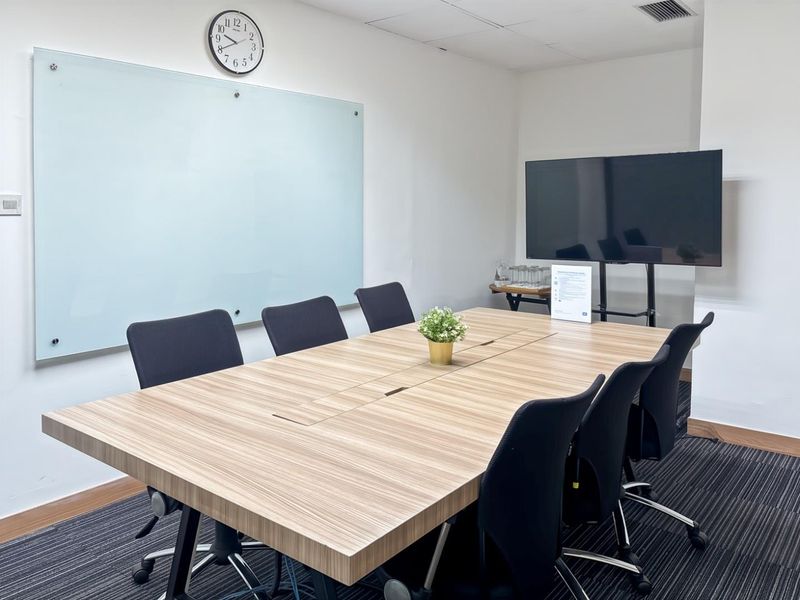 A professional office meeting room featuring a large conference table, wall-mounted glass whiteboard, and carpeted flooring.