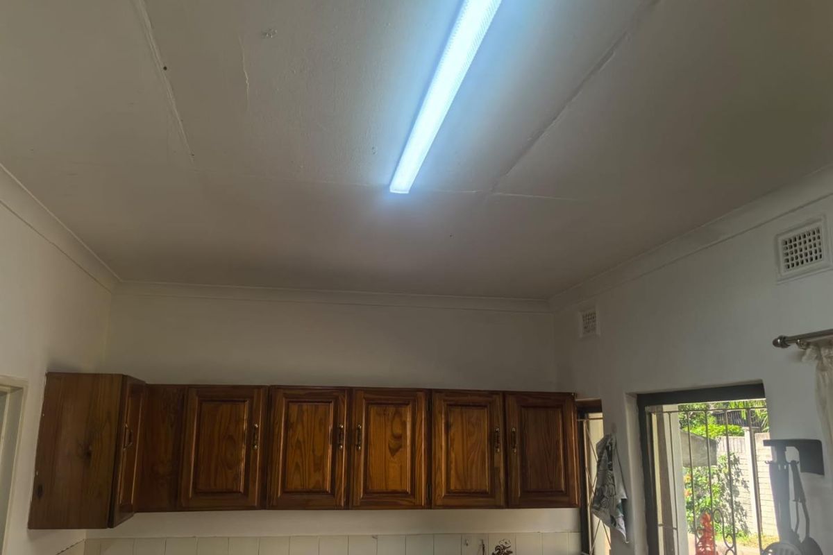 A kitchen area featuring timber cabinetry, tiled splashbacks, and a fluorescent light fixture, showing signs of wear and ceiling plaster cracks.