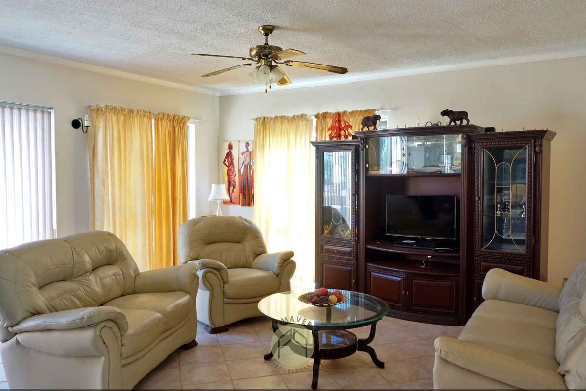 A spacious living room featuring tiled flooring, a ceiling fan, and large windows with vertical blinds, offering a comfortable and functional living space.