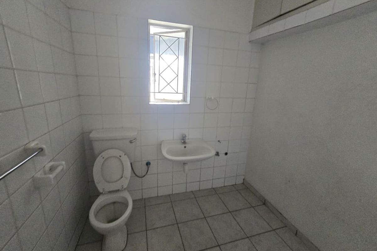 A functional office restroom featuring tiled walls and flooring, a standard toilet, and a wall-mounted basin with security bars on the window.