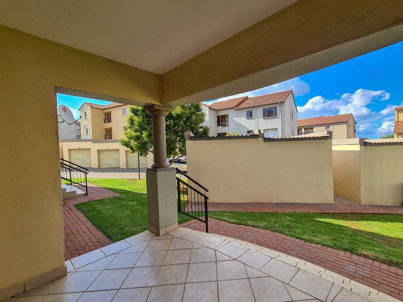 A covered patio area featuring tiled flooring and a structural pillar, overlooking a communal garden and paved walkway within the apartment complex.