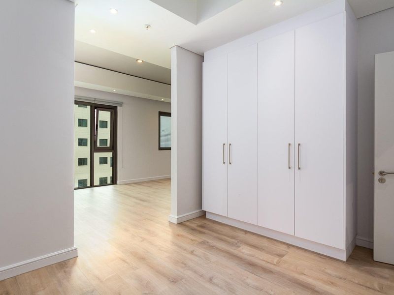 A bright, modern bedroom featuring light wood-look laminate flooring, ample built-in wardrobe storage, and recessed ceiling lighting.