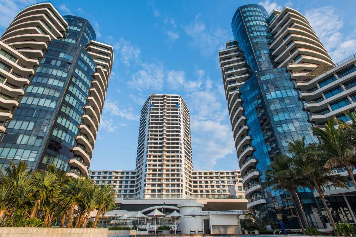 A low-angle view of a modern high-rise apartment complex featuring glass facades, balconies, and a landscaped pool area at the base.