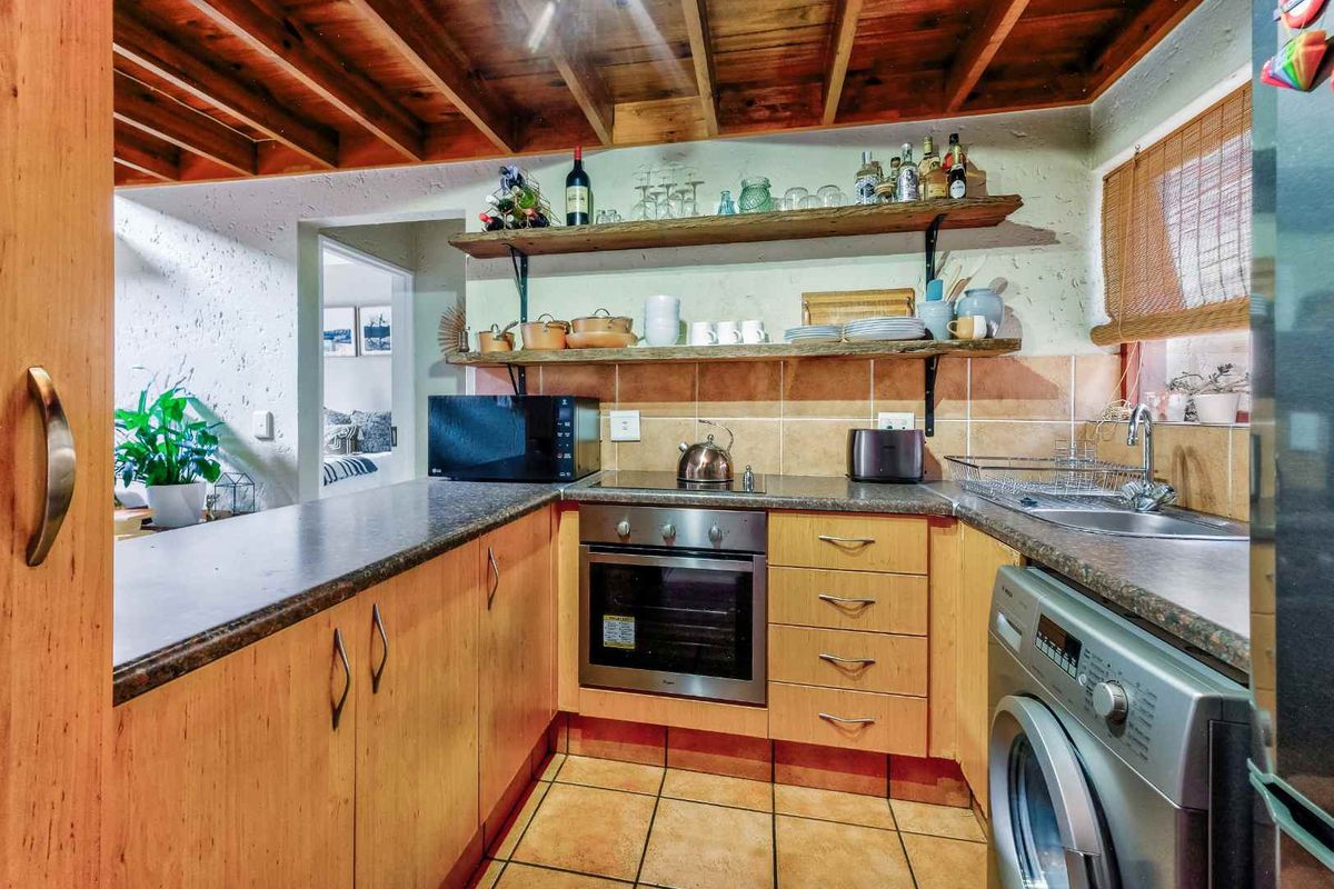 Compact kitchen featuring wooden cabinetry, open shelving, built-in oven, hob, and integrated laundry space with tiled flooring and exposed timber ceiling beams.