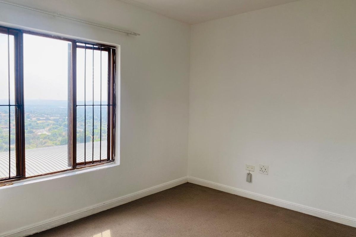 A bright, empty bedroom featuring carpeted flooring, neutral walls, and a window with security bars offering an elevated view.