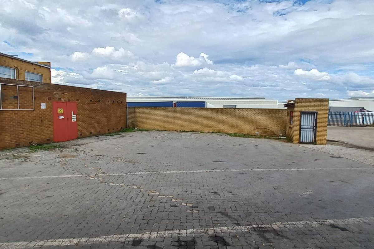 An industrial paved yard area featuring a perimeter brick wall, security gatehouse, and access point, suitable for parking or logistics operations.