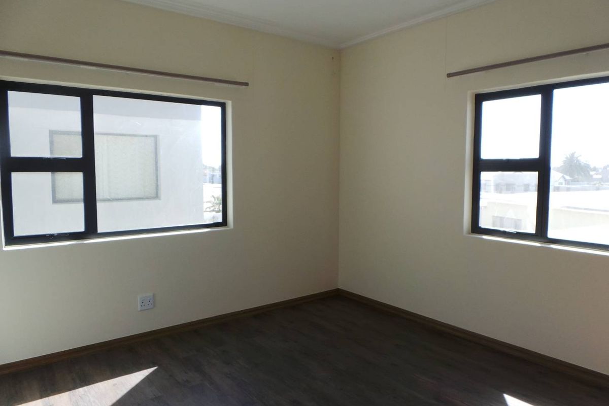 A bright, empty bedroom featuring dark wood-look flooring, neutral walls, and large windows with dark frames.