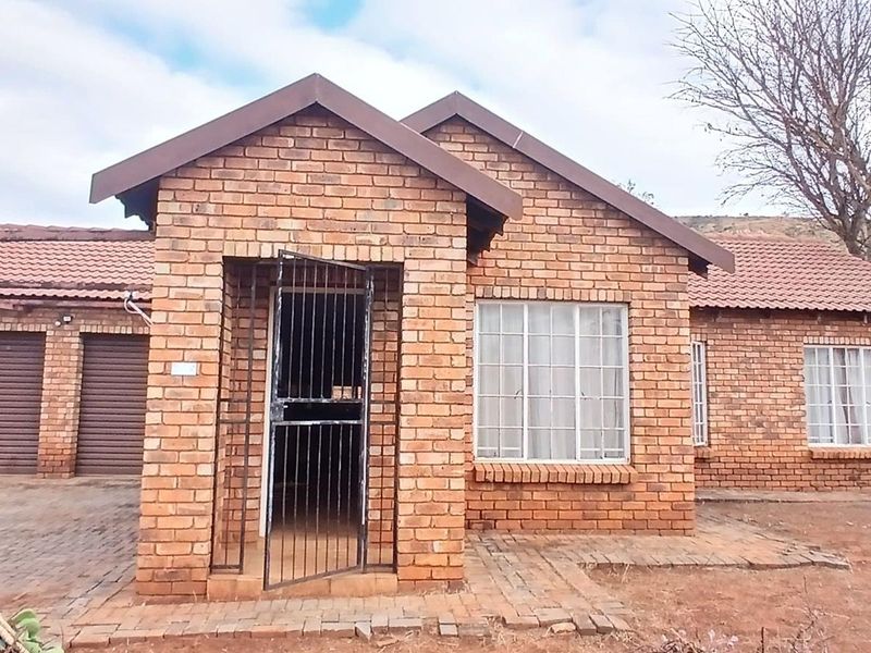 Front view of a face brick townhouse featuring a double garage, tiled roof, and secure entrance gate with paved driveway access.