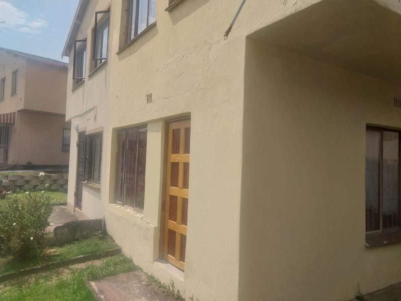 Rear exterior view of a two-story residential building featuring plastered walls, timber door, and metal-framed windows with security bars.