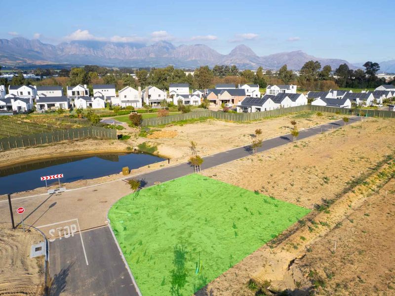 Aerial view of a vacant residential plot within a developed estate, featuring paved road access and proximity to a scenic pond and mountain backdrop.