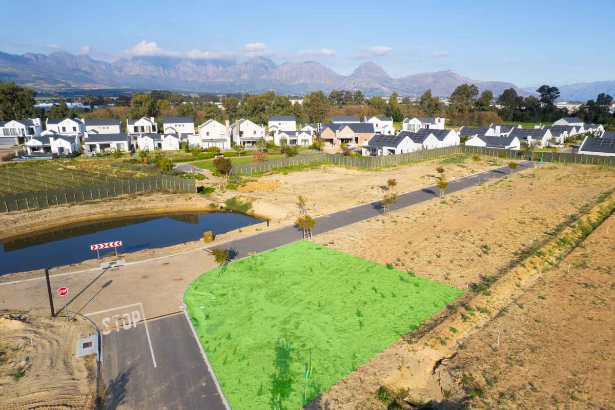 Aerial view of a vacant residential plot within a developed estate, featuring paved road access and proximity to a scenic pond and mountain backdrop.
