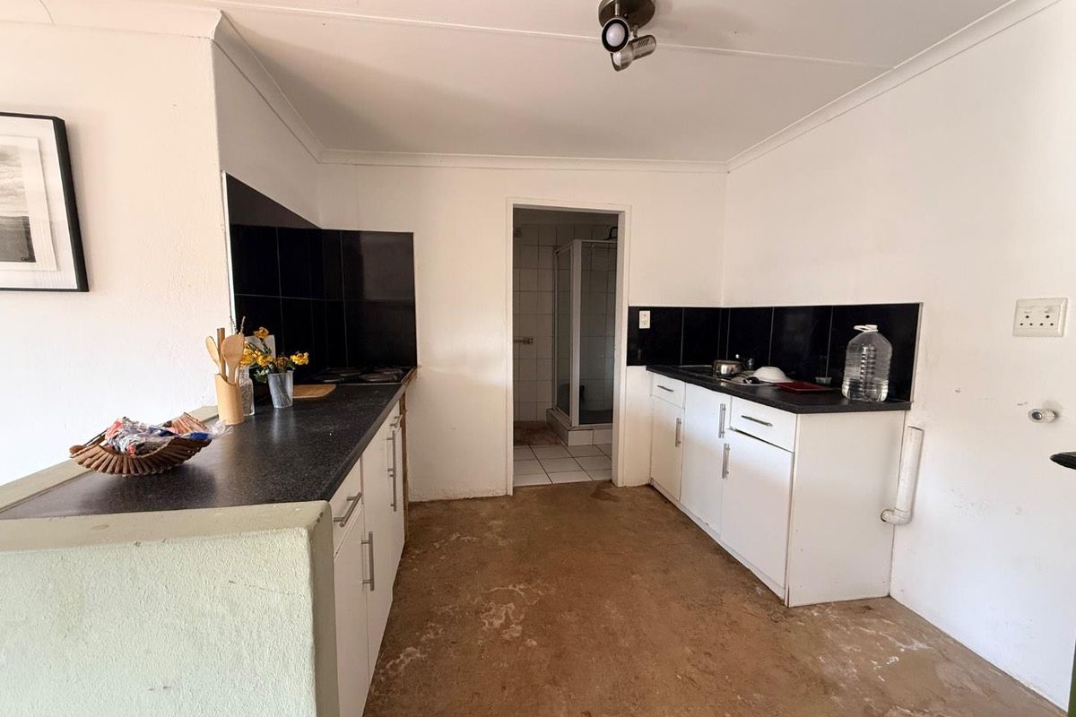 A dated kitchen featuring basic cabinetry, black tiled splashbacks, and a worn floor surface, with an adjacent bathroom visible through the doorway.