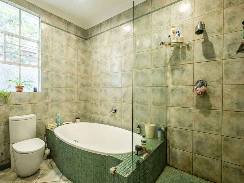 A functional bathroom featuring a built-in bathtub, glass shower screen, and tiled finishes throughout, offering a practical layout with natural light.