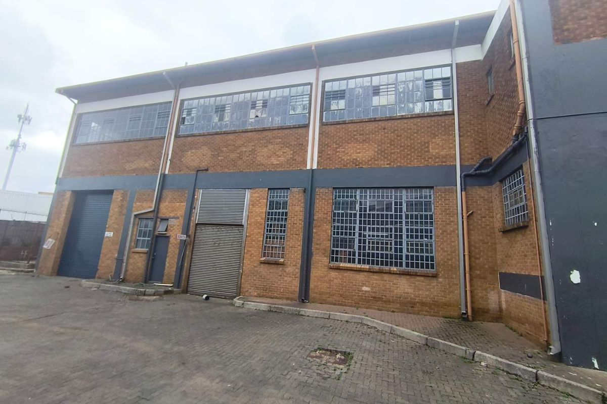 Industrial building exterior featuring brick facade, steel-framed windows, and roller shutter doors with a paved driveway area.
