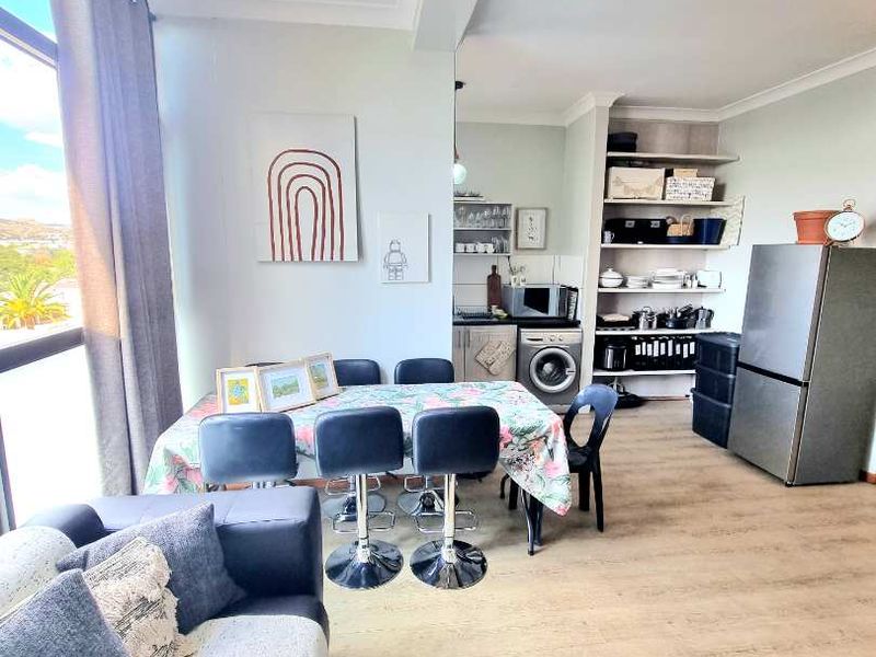 Open-plan living and dining area featuring laminate flooring, large windows, and a compact kitchen space with built-in shelving and cabinetry.