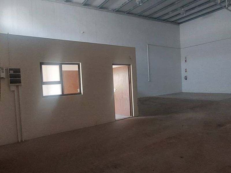 Spacious industrial warehouse unit featuring high ceilings with exposed steel trusses, a roller shutter door, and a partitioned internal office space.