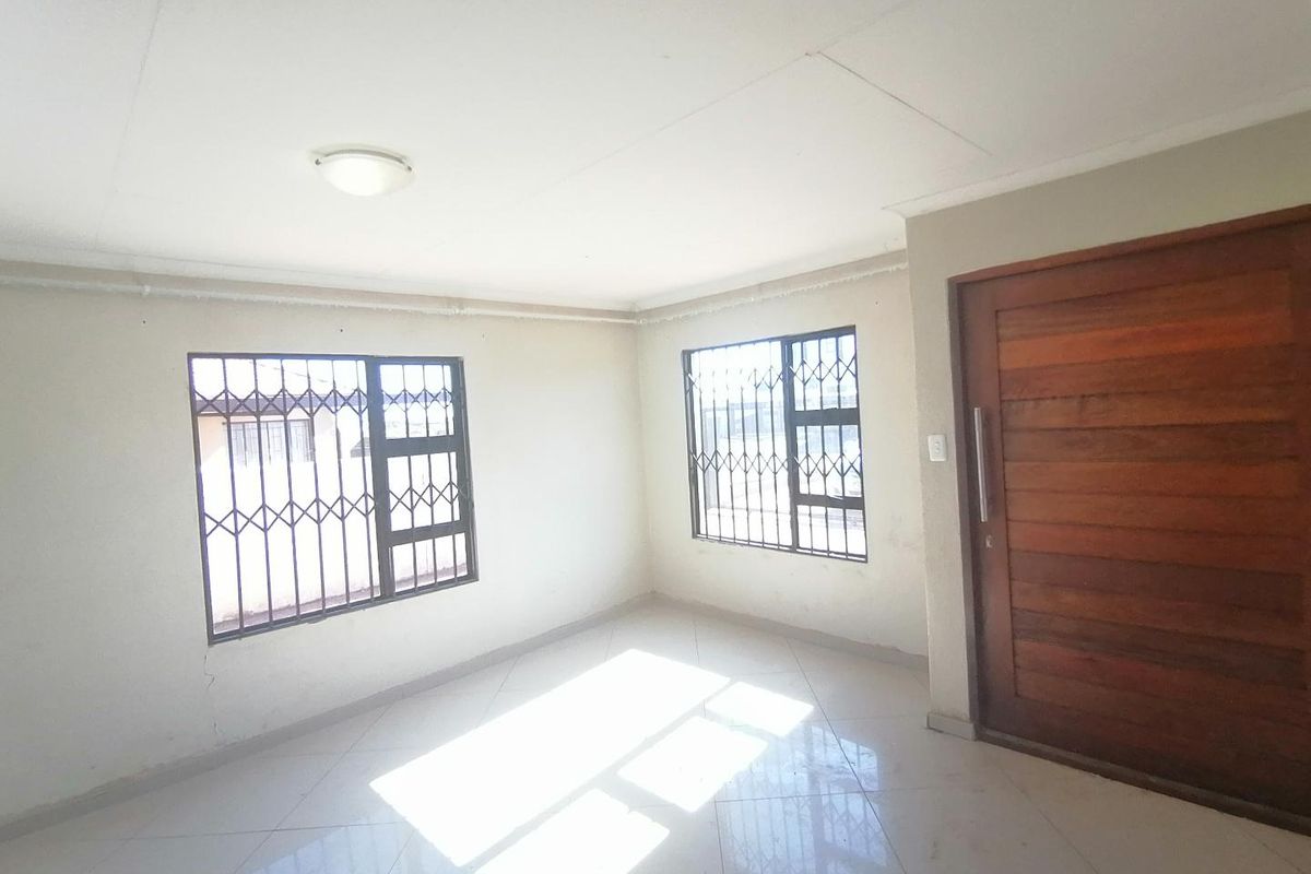 A bright, empty living area featuring tiled flooring, a solid timber entrance door, and windows fitted with security bars.