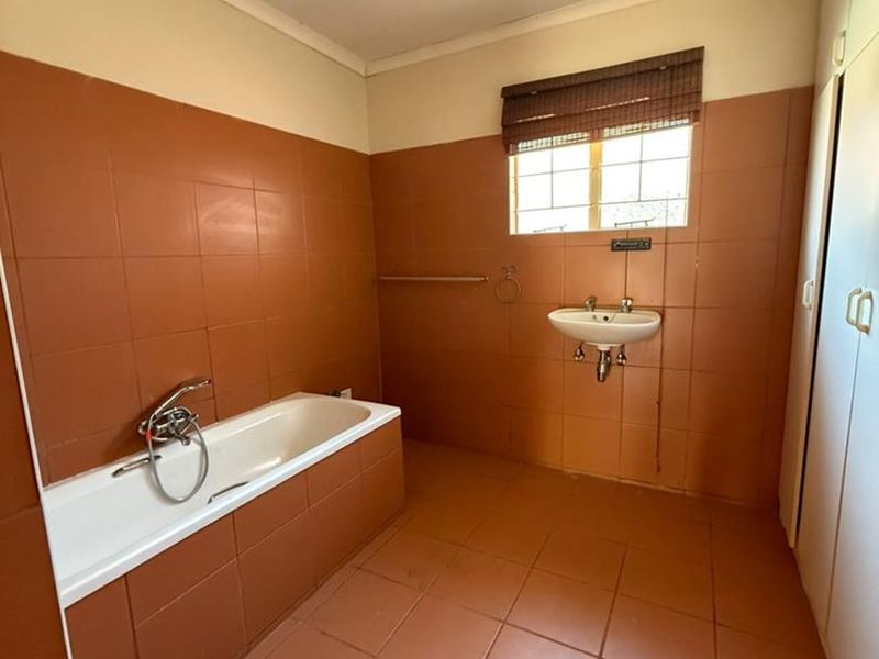A spacious, empty bathroom featuring dated terracotta-style wall and floor tiles, a built-in bathtub, a wall-mounted basin, and integrated storage cupboards.